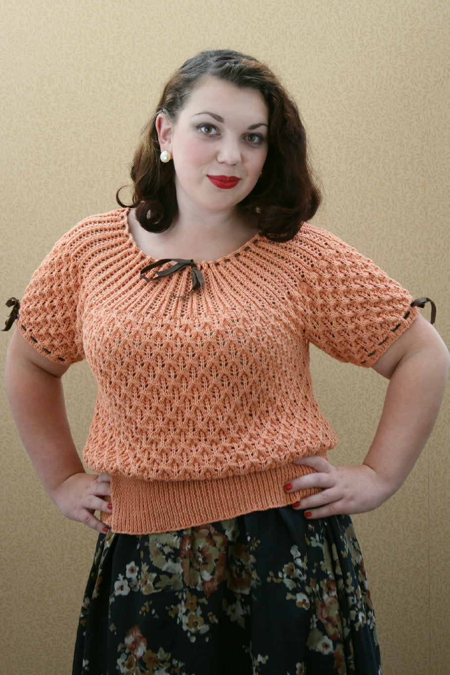 A dark haired woman stands by a neutral background. Her hands are on her hips. She wears pearl earrings, a black floral skirt and an apricot coloured knit top. The top has black ribbon threaded through the neckline and sleeve cuffs. The yoke as a pattern of diagonal stitches leaning in to the gather from the threaded ribbon. The body and sleeves and a criss cross lace pattern over them.