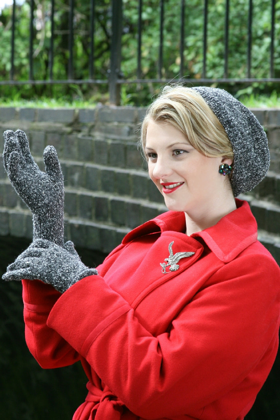 A blonde woman stands outside by a wall and wrought iron fence. She wears a red coat and matching knit hat and gloves. They are knit in a grey and white flecked yarn. The hat sits back on her head so that the front of her hair is visible. She holds one hand up and the other at the wrist of the glove, as if she has just put them on. She also wears a bird brooch.