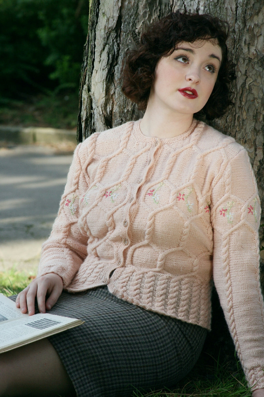A dark haired woman sits on the ground against a tree. She looks upwards and away from the camera with a pensive expression and has a book on her lap. She is wearing a pale pink knit cardigan and a grey and black checked skirt. The Cardigan has an intricate cable pattern over the body and flower decoration across the bustline. 