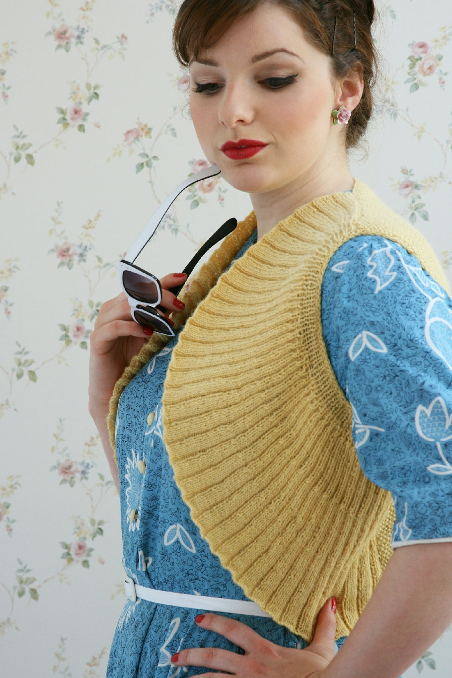 A dark haired woman stands inside with a floral papered wall behind her. Her hair is up and she wears pink flower shaped earrings. She stands sideways on to the camera, one hand on her hip, and the other holding the earpiece of a pair of sunglasses by her mouth. Her eyes are downcast. She is wearing a blue patterned dress with a yellow knit bolero over the top. The bolero is sleeveless and a circular shape which makes it look a little like sun rays.