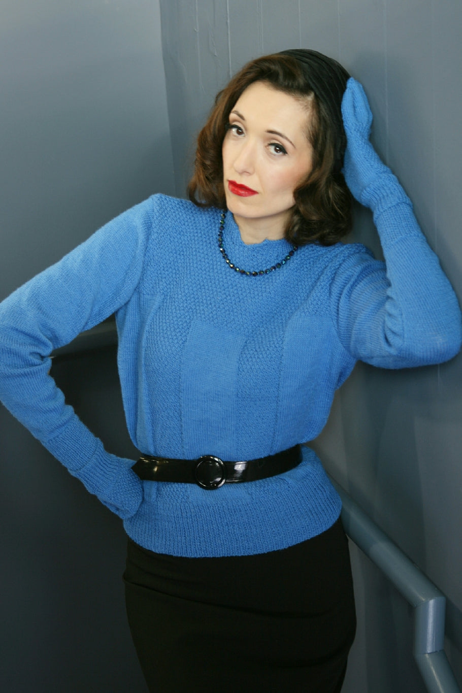 A woman stands in what appears to be a stairwell. She has one hand on her hip and the other on her head, leaning against the wall. She is wearing a blue knit jumper with a textured stitch around the yoke and in panels down the front. She wears a black belt around her middle. She also wears matching gloves.