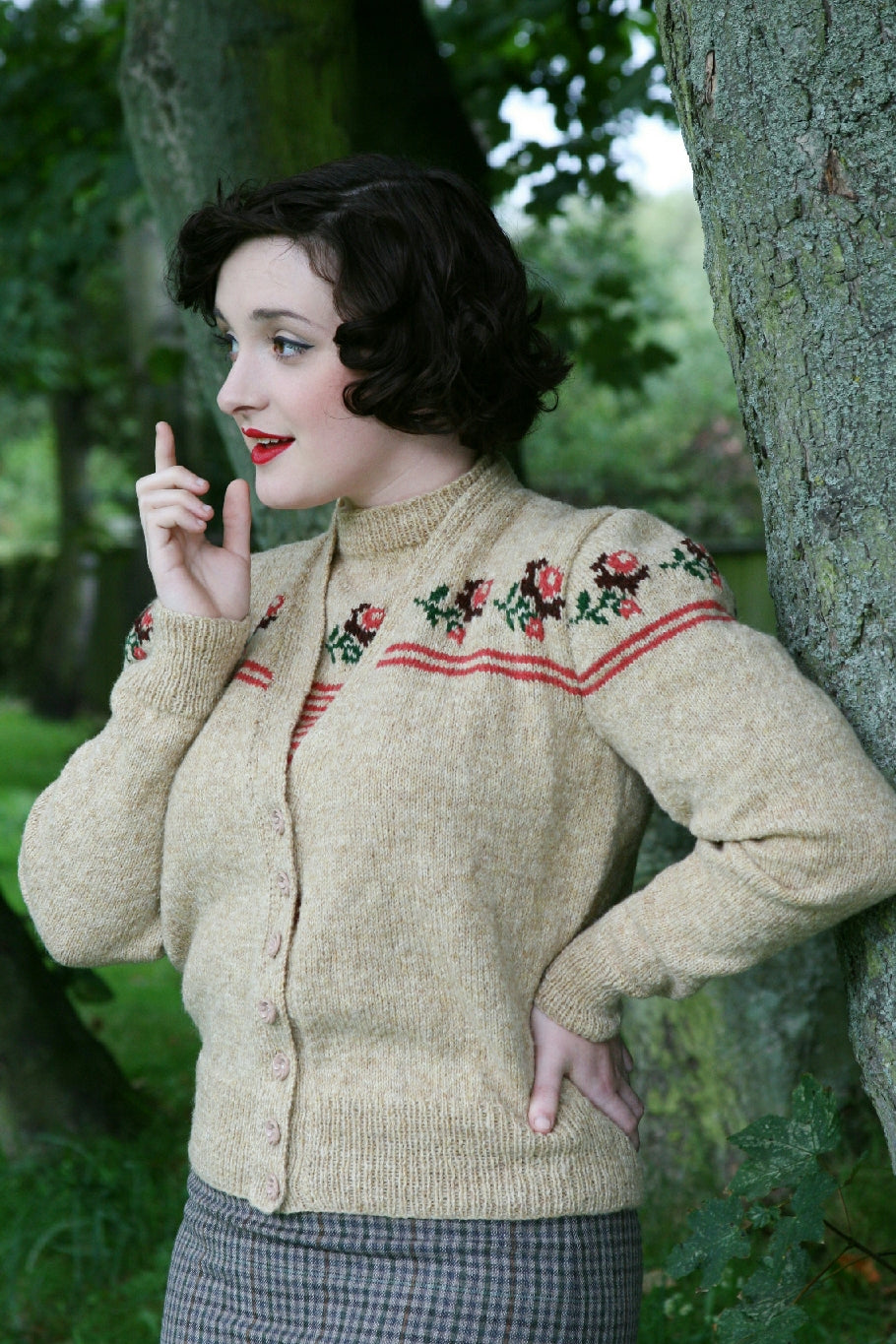 A dark haired woman stands outside by a tree. She has one hand up by her face as if thinking aloud and the other on her hip. She wears a grey, blue and black checked skirt and a matching knit cardigan and top in a beige colour with a decorative pattern of roses around the yoke and top of the sleeve.