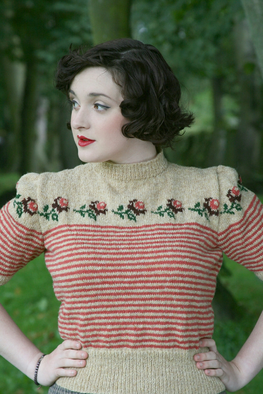A dark haired woman stands outdoors with trees behind her. She looks away from the camera and stands, hands on hips. She is wearing a high necked knit jumper with short sleeves. The yoke is decorated with a pattern of roses. The body and sleeves are striped in red and beige.