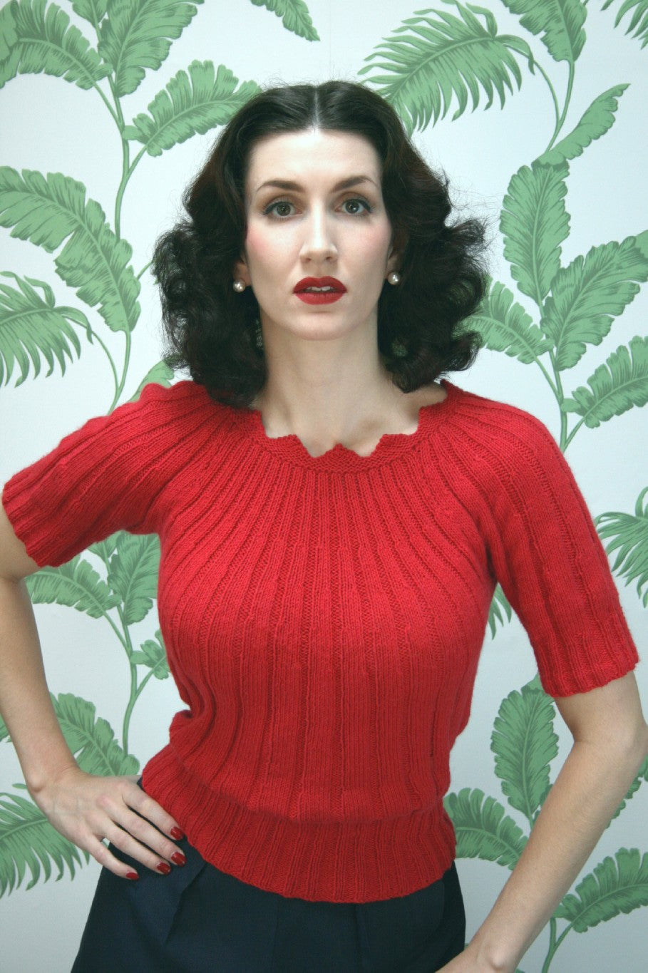 a woman wears a bright red ribbed sweater and matching lipstick. One hand is on her hip and she is looking straight at the camera 