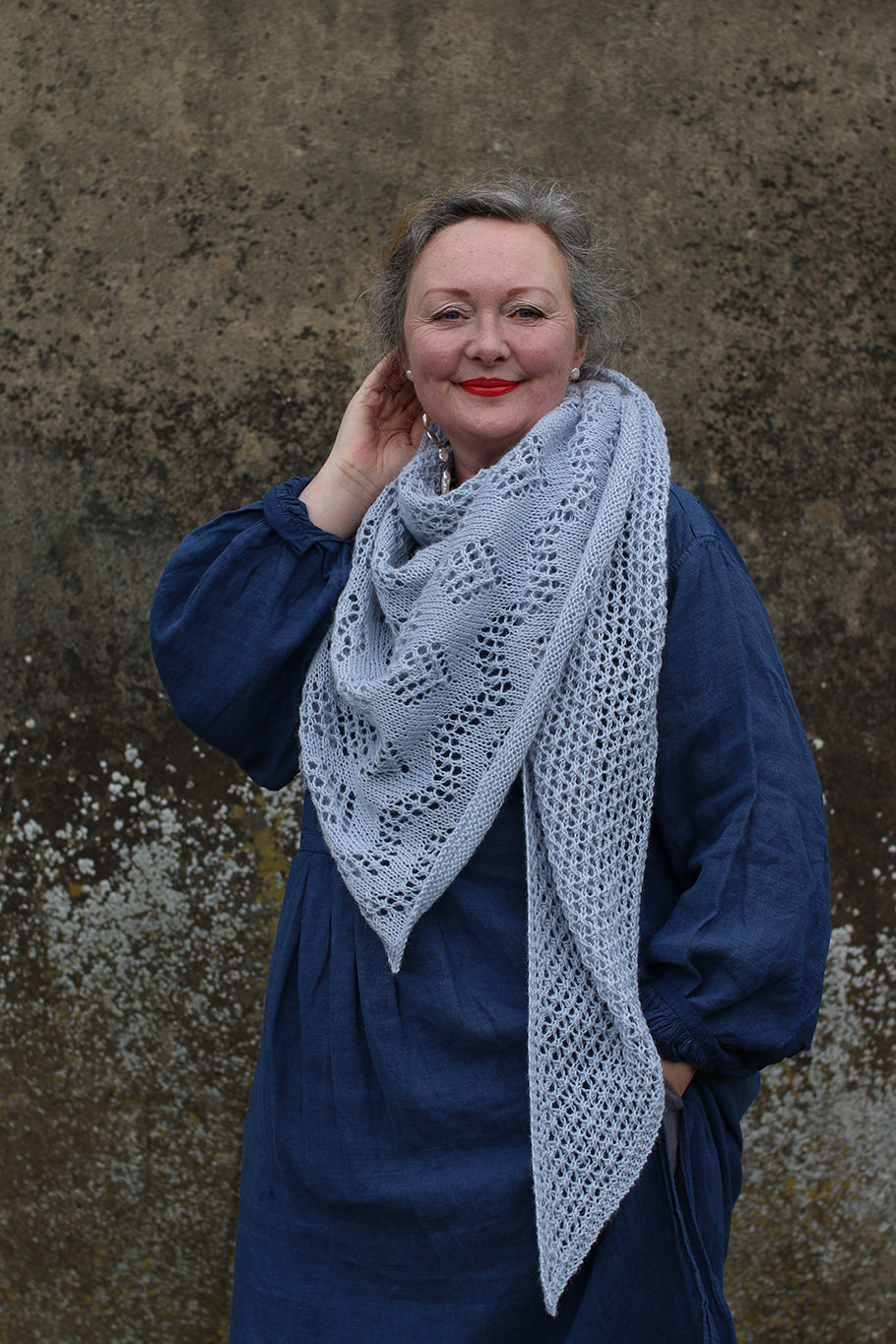 A woman dressed in blue stood in front of a concrete wall. She is dressed in blue with a lace shawl around her neck and shoulders.